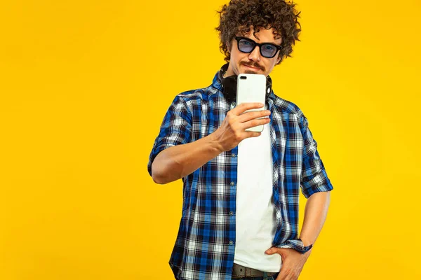 European man with curly hair in blue sunglasses with mobile phone or smarphone. Handsome smiled stylish hipster in plaid shirt posing over yellow background. — ストック写真