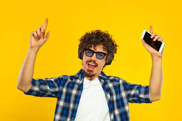 European man with curly hair in blue sunglasses with mobile phone or smarphone. Handsome smiled stylish hipster in plaid shirt posing over yellow background. — 图库照片