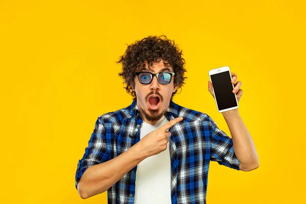青いサングラスで巻き毛を持つヨーロッパの男は、携帯電話やスマートフォンの画面を見て指しています。黄色の背景の上にポーズをとったプレイシャツのハンサムなスタイリッシュなヒップスター. — ストック写真