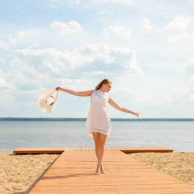 Romantic girl in a white dress with a white hat turned to the long-tailed wooden pier on the beach. clipart