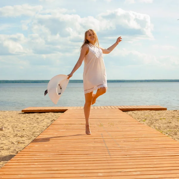 Romantisches Mädchen in weißem Kleid mit weißem Hut, das sich dem langschwänzigen Holzsteg am Strand zuwendet. — Stockfoto