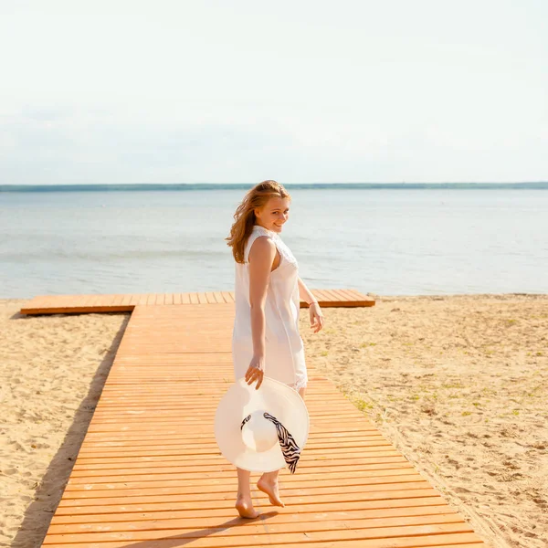 Romantická dívka v bílých šatech s bílým kloboukem se obrátil k dlouho sledoval dřevěné molo na pláži. — Stock fotografie