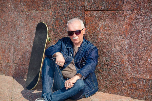 Äldre man sitter och drömmer på trottoaren nära en skateboard Stockbild