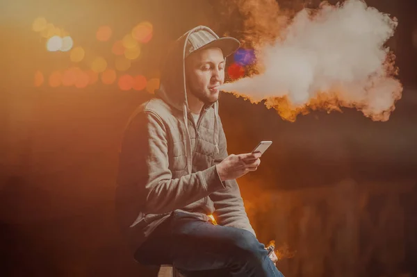 Jovem fumando um cigarro eletrônico olhando para o telefone — Fotografia de Stock