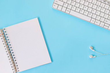 Mavi kağıt arka planda dizüstü bilgisayarı, defteri ve kulaklığı olan beyaz ofis masasının üst görüntüsü.