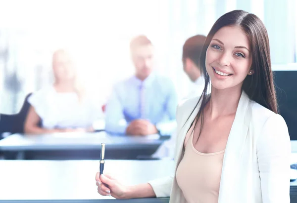 Lächelnde selbstbewusste Geschäftsfrau, die mit ihren Kollegen im Hintergrund im Büro in die Kamera schaut — Stockfoto