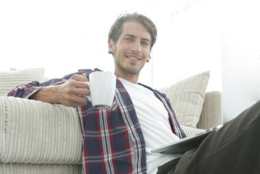 genç adam kanepede yakın katta oturan bir fincan tutan dizüstü bilgisayar ile