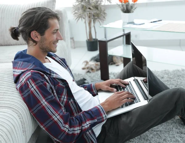现代的家伙坐在沙发旁边的地板上的一台笔记本电脑 — 图库照片