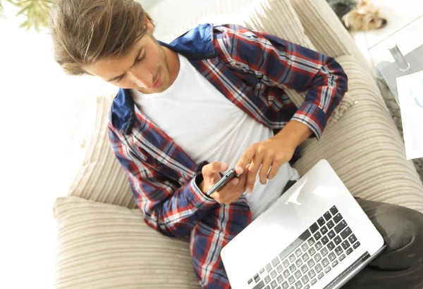 Jeune homme sérieux avec ordinateur portable et smartphone dans le salon . — Photo