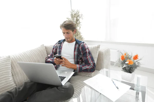 Jeune homme sérieux avec ordinateur portable et smartphone dans le salon . — Photo