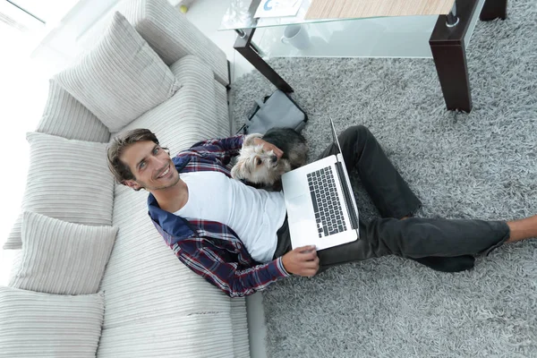 Guapo chico con portátil abrazando a su perro y sentado cerca del sofá — Foto de Stock