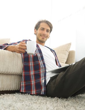 genç adam kanepede yakın katta oturan bir fincan tutan dizüstü bilgisayar ile