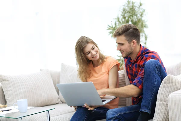 Jovem casal está usando um laptop e sorrindo enquanto sentado no sofá em casa — Fotografia de Stock