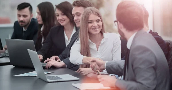 Grupo de gente de negocios confiados en formalwear sentado a la mesa juntos y sonriendo mientras dos hombres apretón de manos —  Fotos de Stock