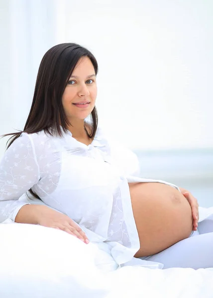 Beautiful pregnant woman sitting at sofa and keeping hand on belly. — Stock Photo, Image