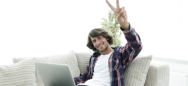 Щасливий хлопець з ноутбуком сидить на дивані і показує йому руку виграшний жест — стокове фото