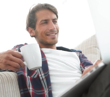 genç adam kanepede yakın katta oturan bir fincan tutan dizüstü bilgisayar ile