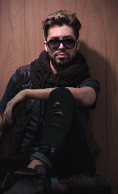 Güneş gözlüklü rahat moda adamı stüdyoda tahtaya oturur.