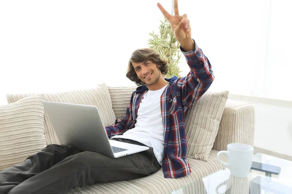 快乐的人，与笔记本电脑坐在沙发上，显示他的手一个胜利的手势 — 图库照片