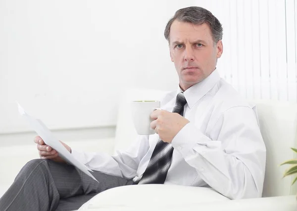 Hombre de negocios confiado bebiendo té y leyendo un documento, sentado en una silla en la habitación del hotel — Foto de Stock