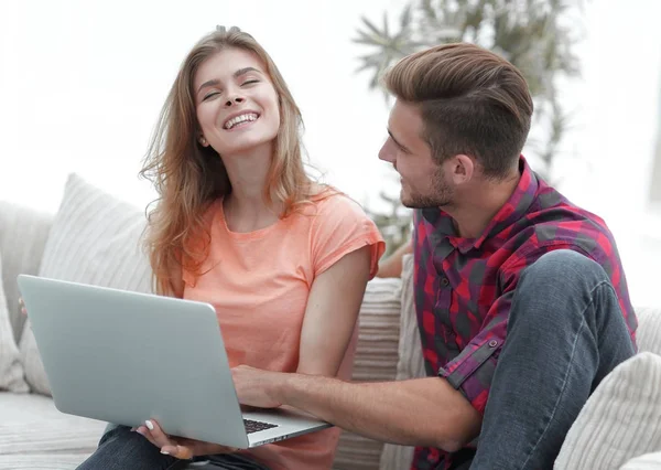 Jovem casal está usando um laptop e sorrindo enquanto sentado no sofá em casa — Fotografia de Stock