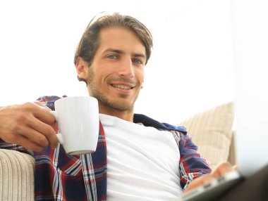 genç adam kanepede yakın katta oturan bir fincan tutan dizüstü bilgisayar ile