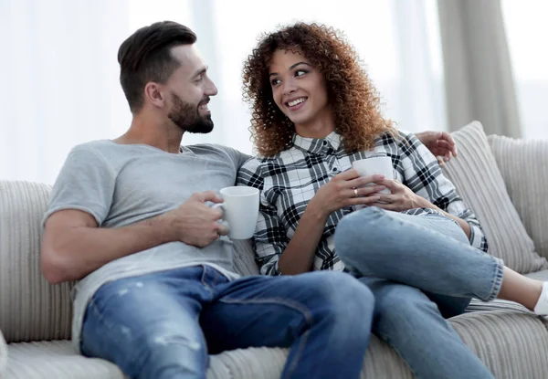 Hermosa pareja sosteniendo una taza de bebida mientras está sentada en el sofá en casa — Foto de Stock