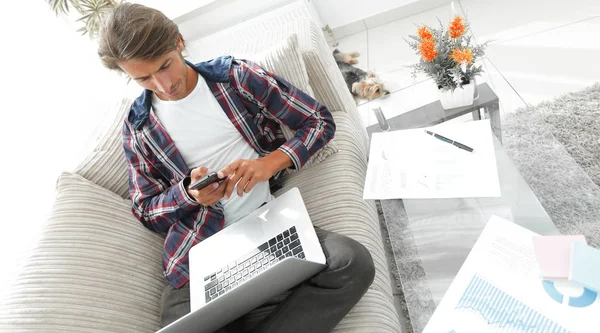 Joven serio con ordenador portátil y teléfono inteligente en la sala de estar . —  Fotos de Stock