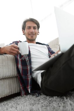 genç adam kanepede yakın katta oturan bir fincan tutan dizüstü bilgisayar ile