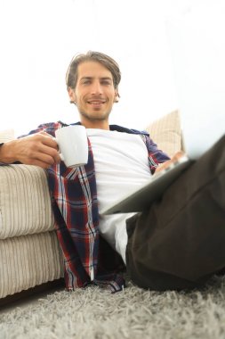 genç adam kanepede yakın katta oturan bir fincan tutan dizüstü bilgisayar ile