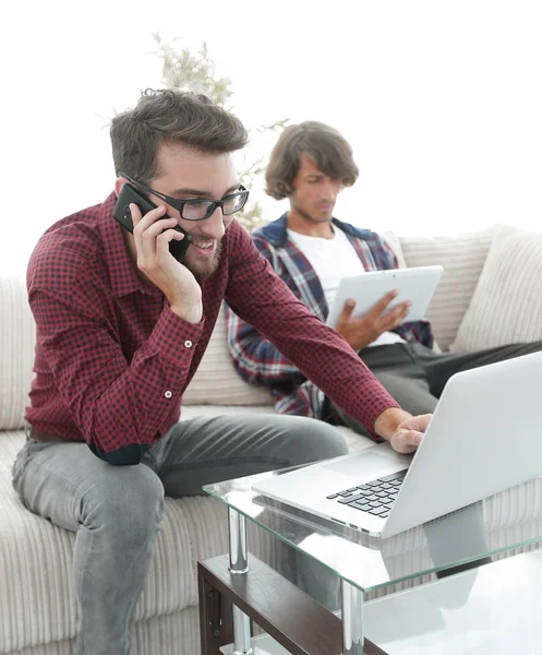 Tipo trabajando en el ordenador portátil y hablando en el teléfono inteligente sentado en el sofá — Foto de Stock