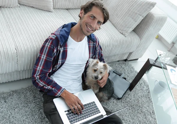 Guapo chico con portátil abrazando a su perro y sentado cerca del sofá —  Fotos de Stock