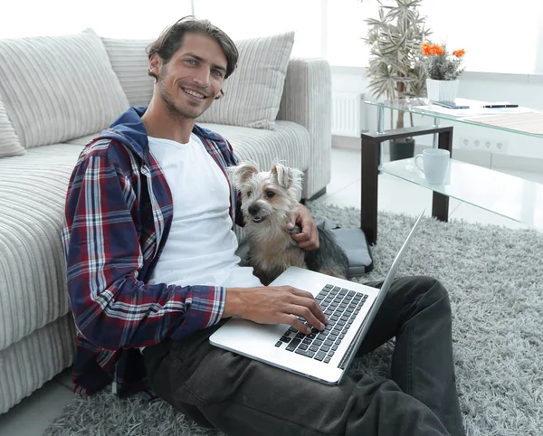 Guapo chico con portátil abrazando a su perro y sentado cerca del sofá — Foto de Stock