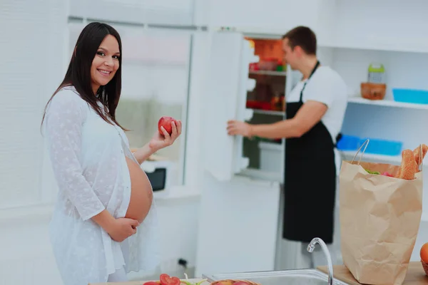 Zwangere vrouw met een appel op de achtergrond van haar echtgenoot opende de koelkast. — Stockfoto