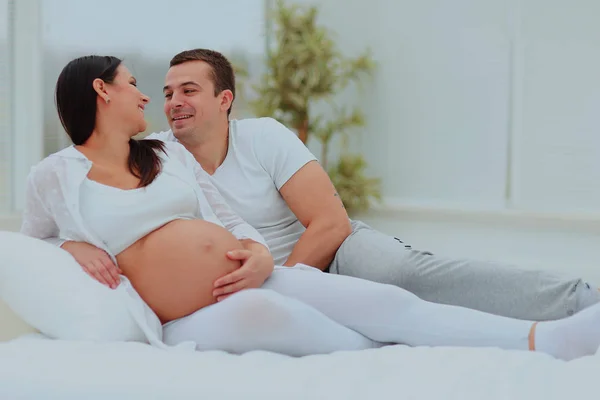 Člověk vypadá láskyplně na svou těhotnou ženu ležící na bílé posteli. — Stock fotografie