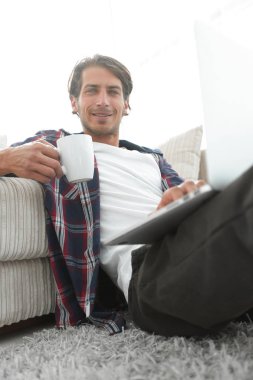 genç adam kanepede yakın katta oturan bir fincan tutan dizüstü bilgisayar ile