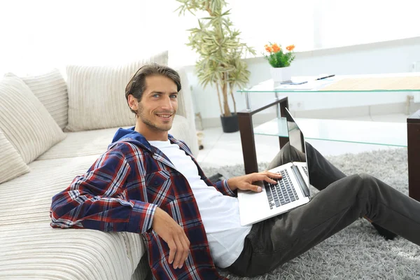 Jovem sorridente com laptop sentado no chão perto do sofá — Fotografia de Stock