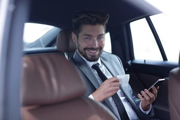 Uomo d'affari che beve caffè in auto con il telefono in mano — Foto Stock