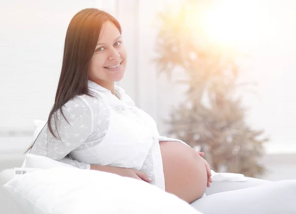 Mulher grávida bonita sentada no sofá e mantendo a mão na barriga . — Fotografia de Stock