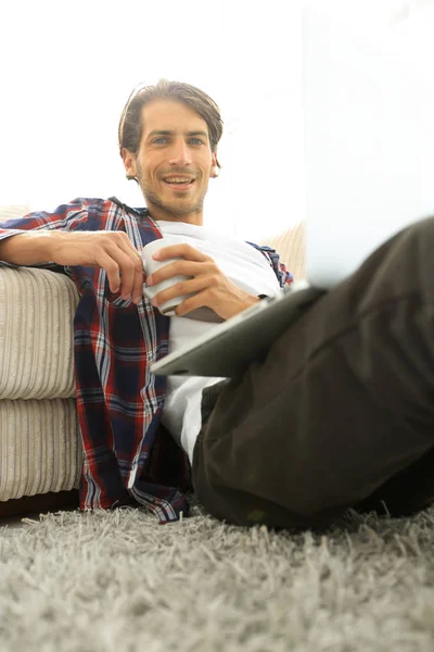 Joven con portátil sosteniendo una taza sentada en el suelo cerca del sofá —  Fotos de Stock