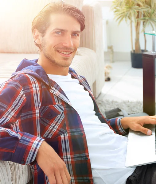 Jovem sorridente com laptop sentado no chão perto do sofá — Fotografia de Stock