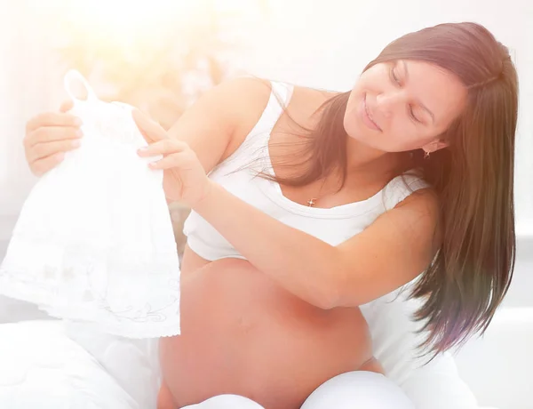 Zwangere vrouw overweegt kinderen shirt, zitten op het bed. — Stockfoto