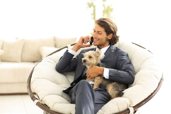 Homme d'affaires tient son animal de compagnie et parle sur le smartphone tout en étant assis dans une chaise confortable — Photo