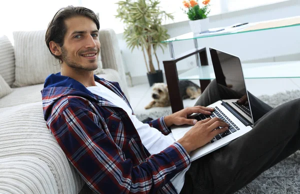 Ragazzo sorridente con un computer portatile seduto sul pavimento vicino al divano — Foto Stock
