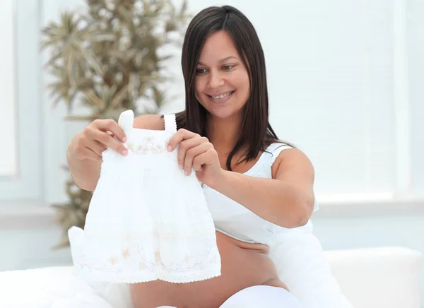Mujer embarazada teniendo en cuenta la camisa de los niños, sentado en la cama . — Foto de Stock