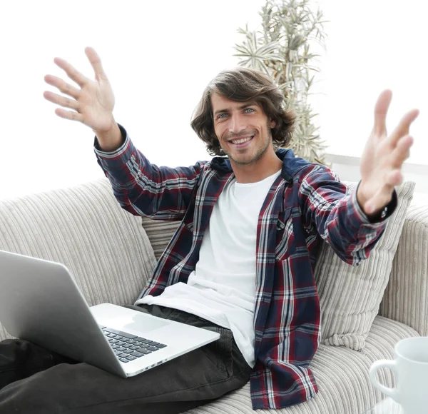 Glücklicher Kerl mit Laptop, der auf dem Sofa sitzt und mit ausgestreckten Händen grüßt. — Stockfoto