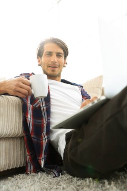 genç adam kanepede yakın katta oturan bir fincan tutan dizüstü bilgisayar ile