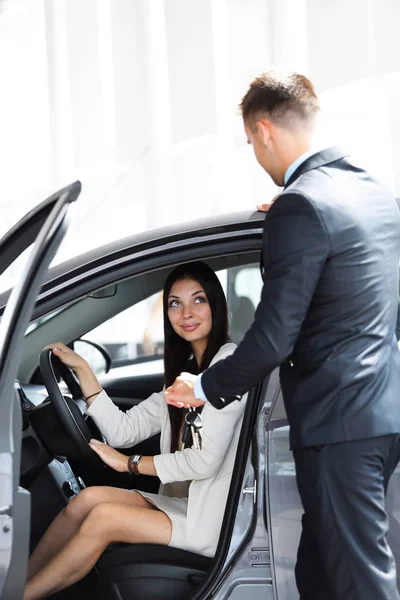 Joven mujer consigue la llave y sonriendo, sentado en un coche nuevo en la sala de exposición . — Foto de Stock