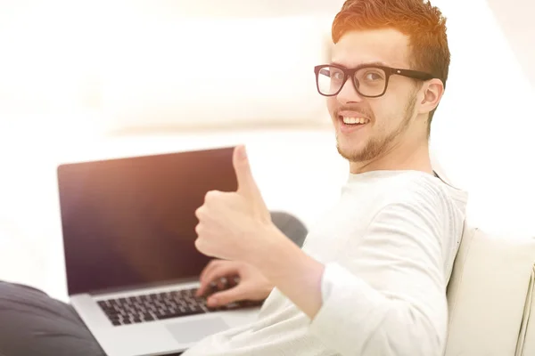 Jovem moderno sentado na frente do laptop aberto e mostrando o polegar para cima — Fotografia de Stock