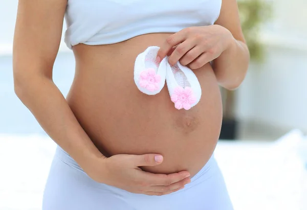 妊娠中の女性は、赤ちゃんの靴を保持しています。彼女の腹部に近い. — ストック写真